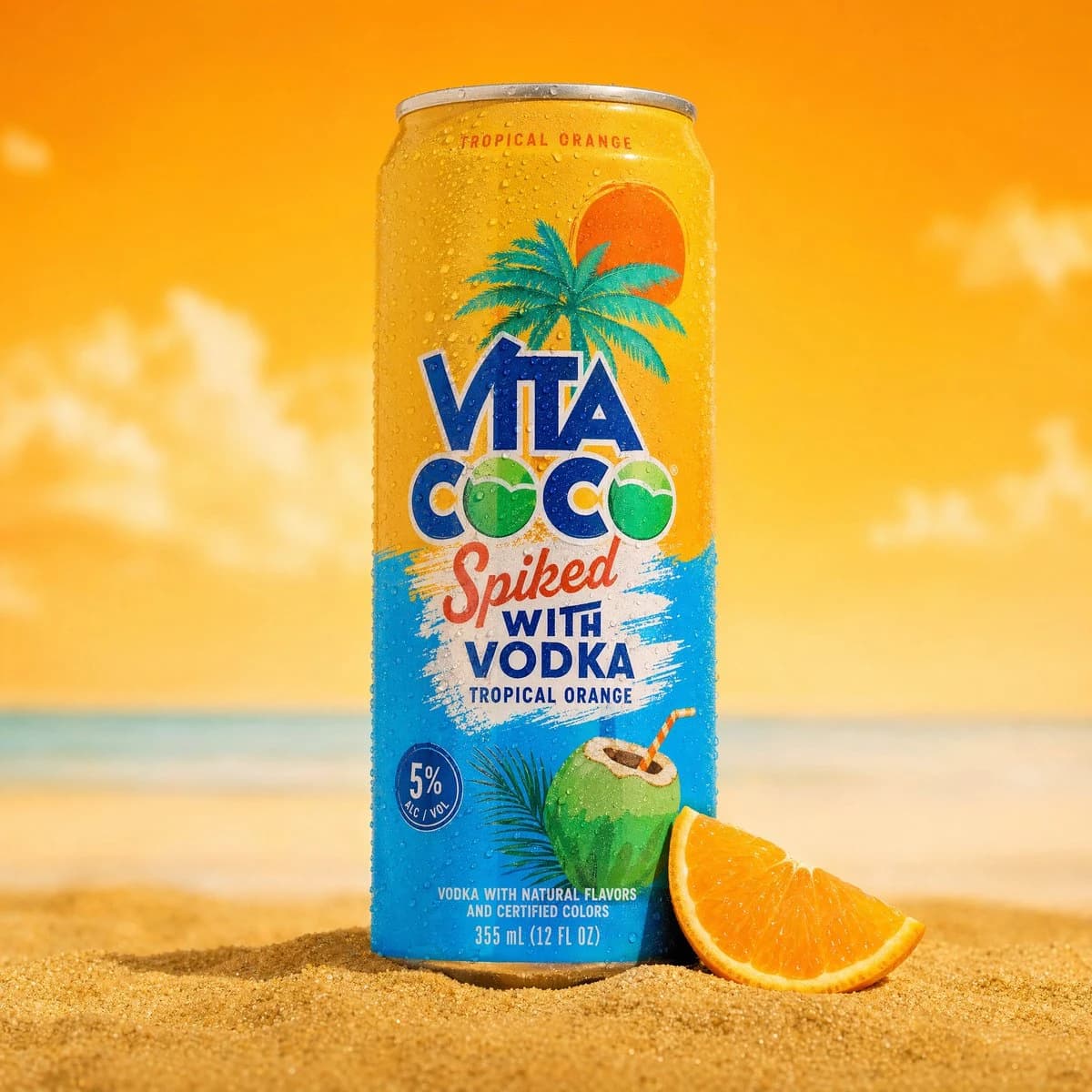 A yellow and blue can of Vita Coco spiked tropical orange drink rests in sand with an orange slice.