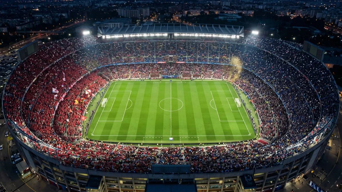 Aerial night view of a packed soccer stadium with bright floodlights, green pitch and red-blue crowds
