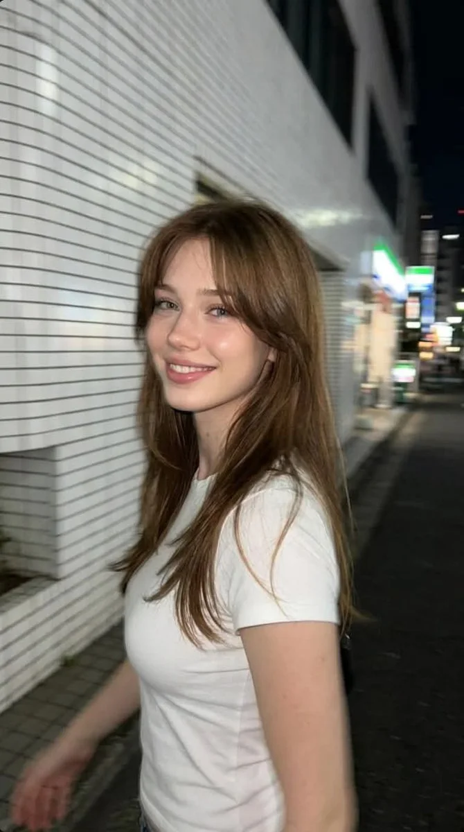 A young woman with long chestnut-brown hair smiles while walking past white ceramic tile walls on a quiet Tokyo sidewalk at night.