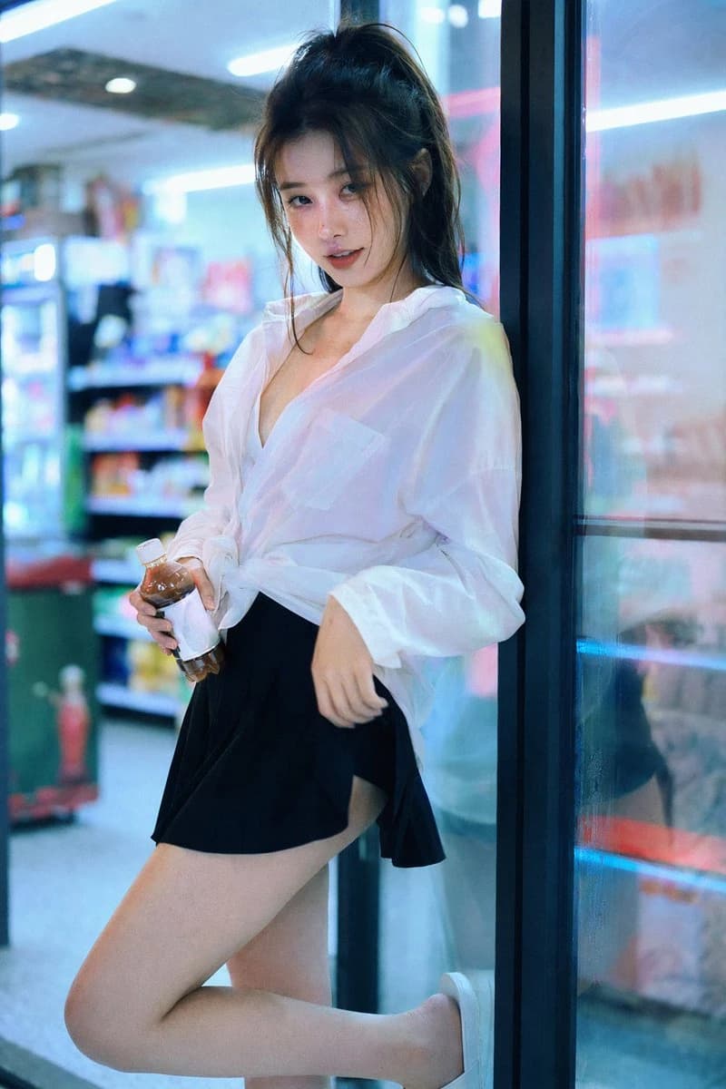 A young woman with delicate features poses in a convenience store, wearing an oversized white shirt and a black mini skirt, illuminated by neon lights