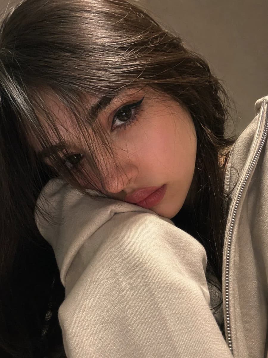Close-up selfie-style portrait of a young woman in a cream hoodie with curtain bangs and soft warm lighting