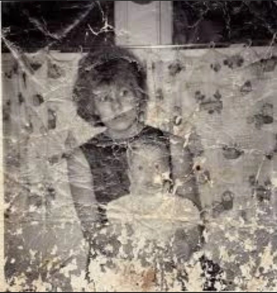 A heavily damaged black-and-white portrait shows a woman in a dark dress holding a toddler against her chest, with a patterned curtain in the background