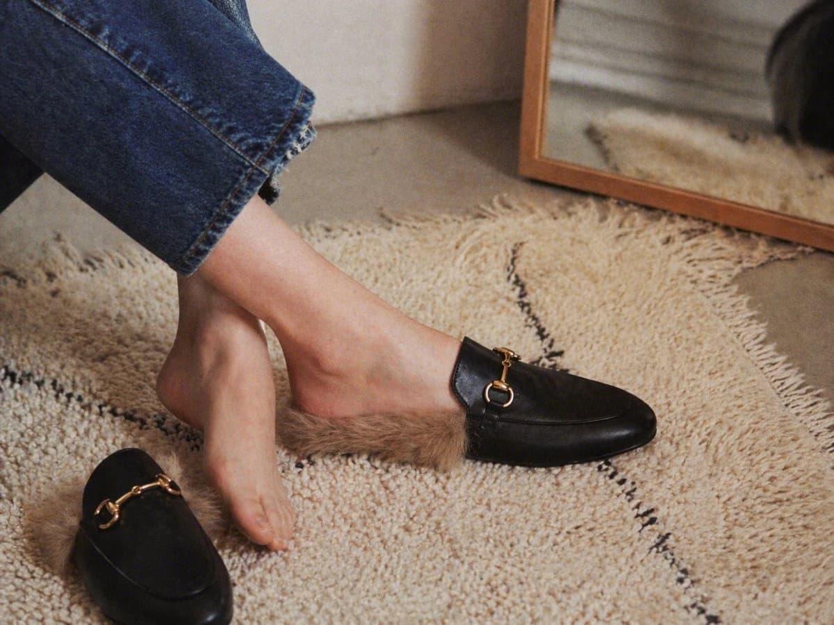 A woman's lower legs and feet showcase black leather backless loafers with tan faux-fur lining on a textured cream shag rug in a cozy interior
