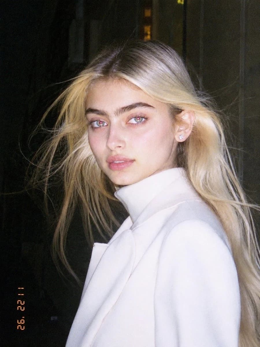 A young woman with long blonde hair poses in a white coat, captured with harsh flash highlights and a nostalgic, low-resolution aesthetic