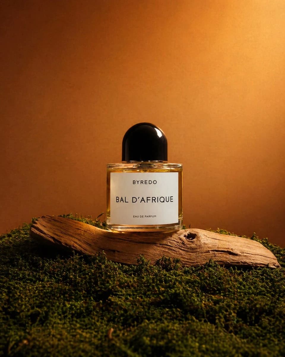 A luxury perfume bottle of BYREDO's BAL D’AFRIQUE is elegantly displayed on driftwood amidst rich green moss, illuminated by warm light
