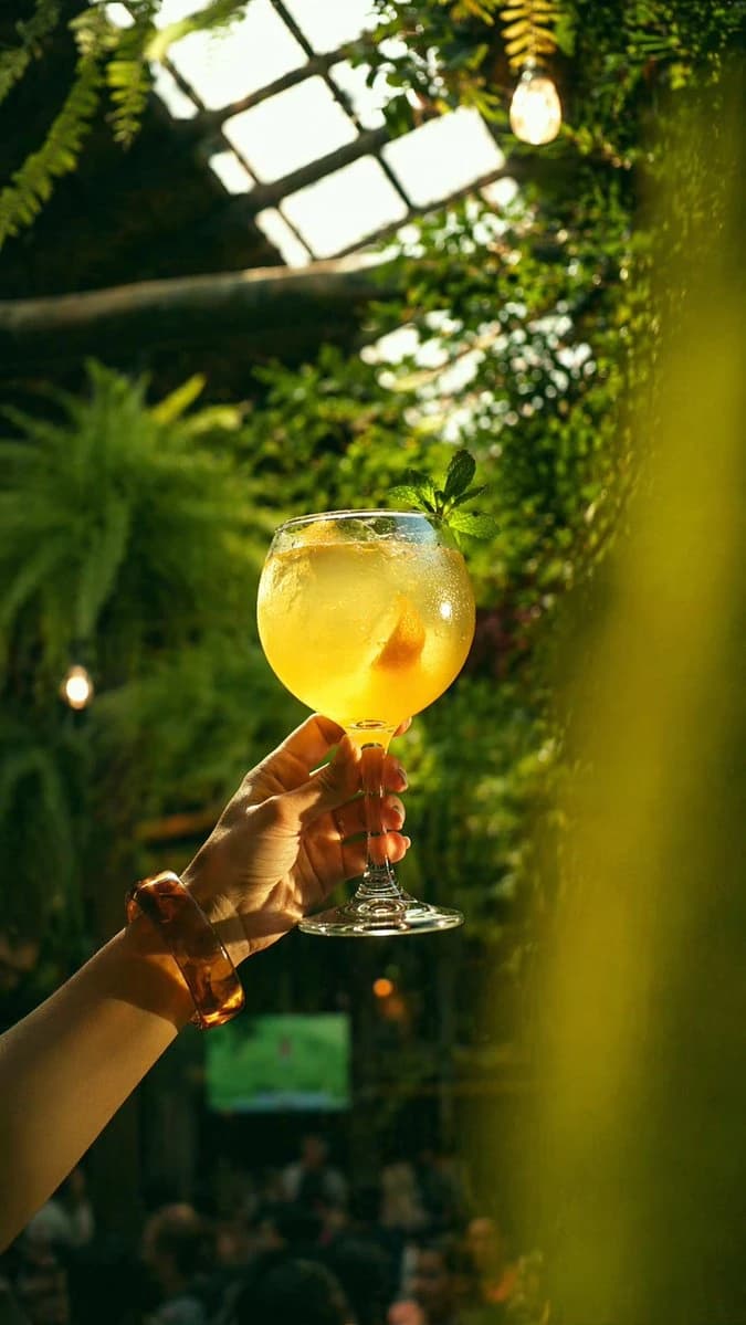 A hand holds a large balloon wine glass filled with a golden citrus cocktail, surrounded by lush greenery in a bright greenhouse bar