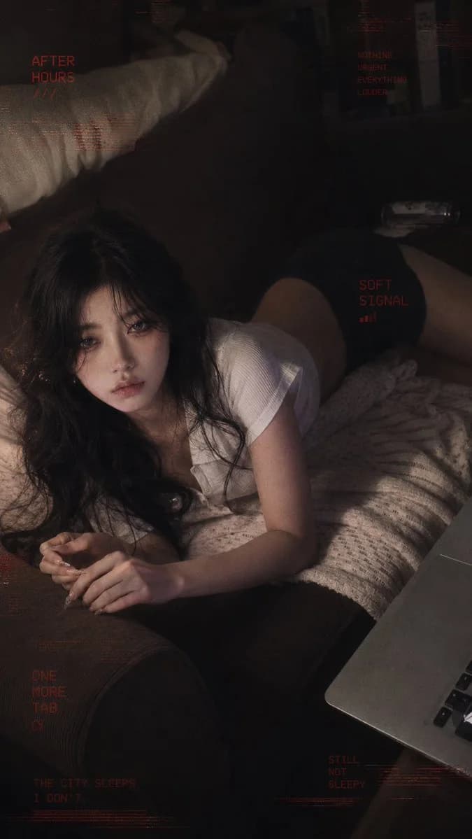 A person with long, wavy black hair lies on a couch, gazing thoughtfully with a soft expression, surrounded by a cozy atmosphere.
