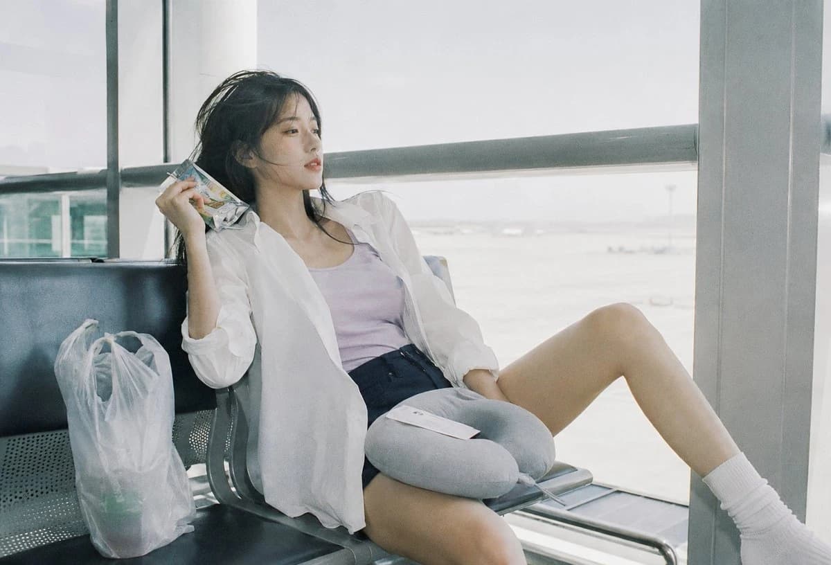 A young woman in a white shirt and denim shorts sits relaxed on an airport bench, holding a snack while gazing out at the runway