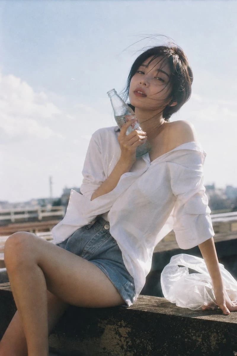 A young woman in an oversized white shirt and high-waisted shorts sits on a rooftop, holding a cold drink as her hair gently blows in the wind