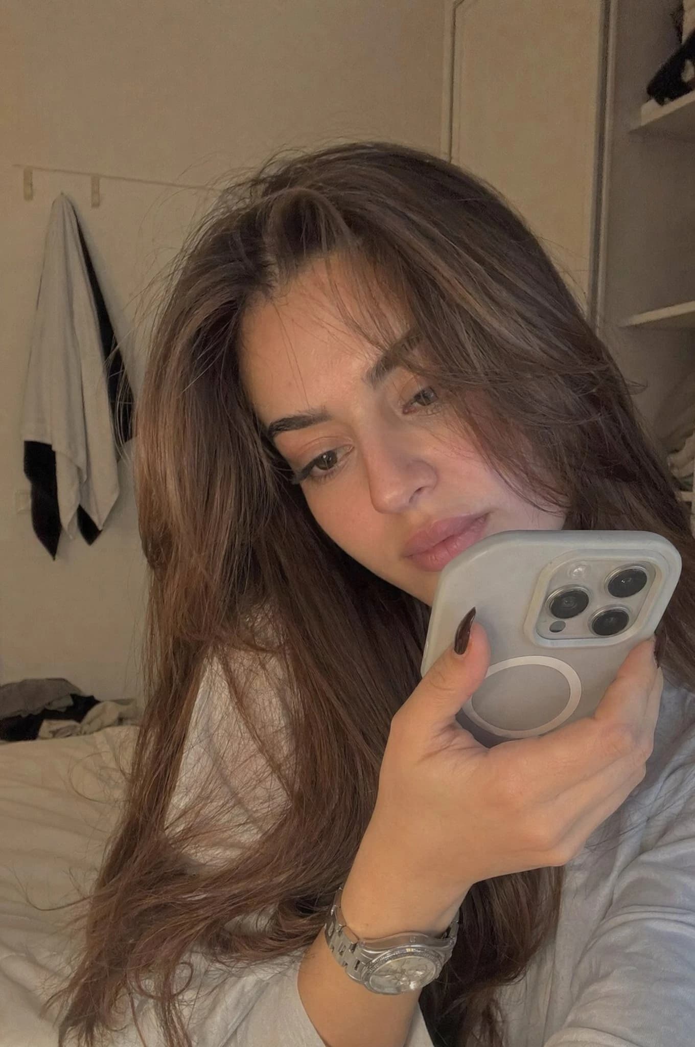 A young woman with long hair sits in a softly lit bedroom, holding her phone close as she records a private video, exuding a thoughtful expression