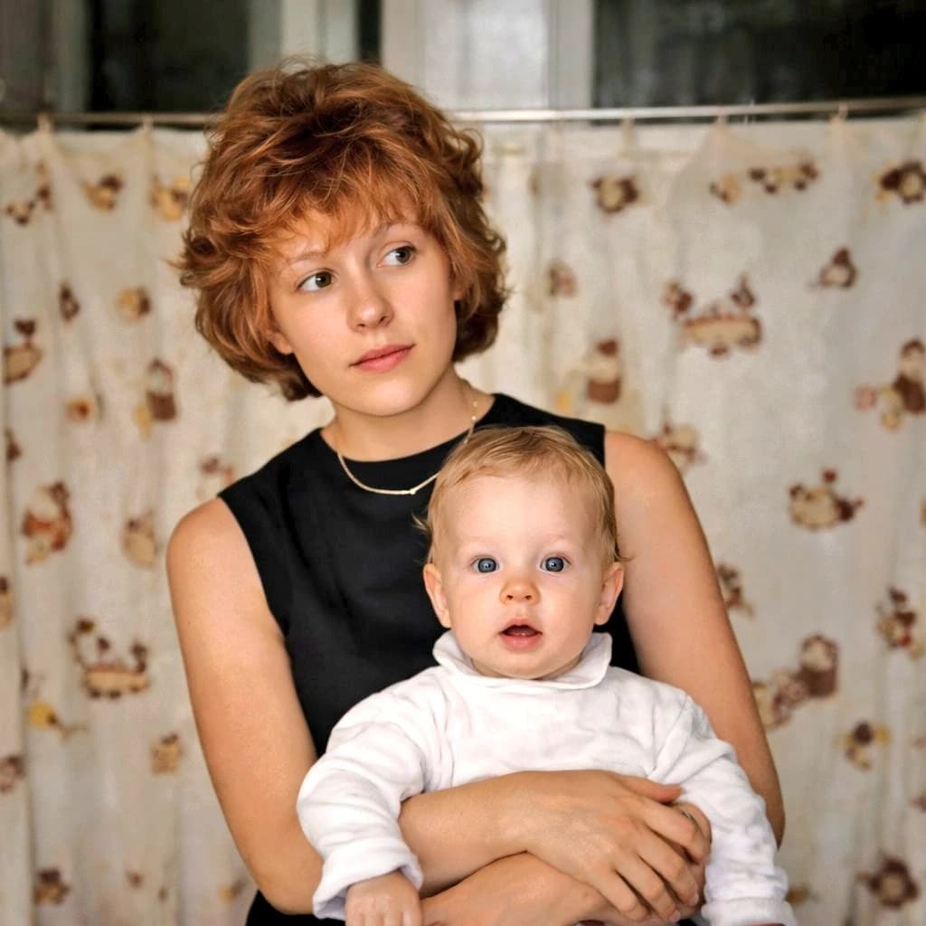 A young mother with auburn hair in a black dress holds her light-haired toddler against a cream curtain with teddy bear motifs, evoking nostalgia