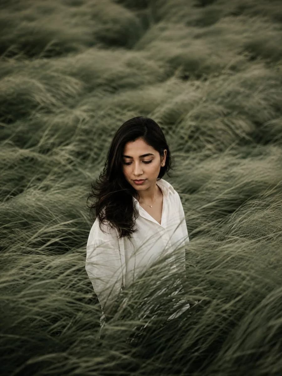 Soft editorial portrait shows a woman in a white blouse standing among tall sage-green grasses