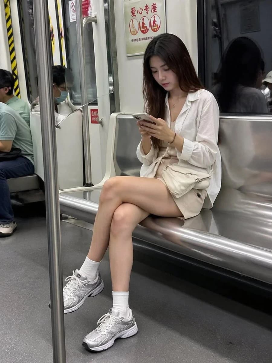 A woman in a white blouse and shorts sits on a subway bench, focused on her phone, with fellow passengers in the background
