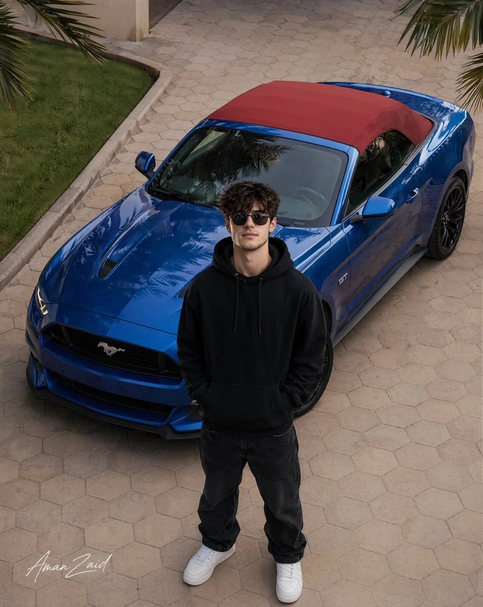 A confident young man in a black hoodie stands near a blue 2017 Ford Mustang GT Convertible with a red soft top, captured from above