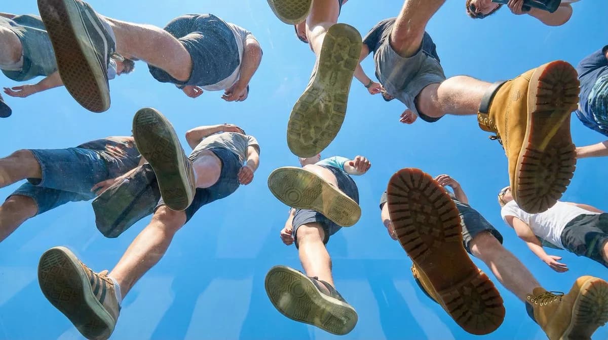 Worm's-eye view through a glass floor shows pedestrians' shoes and legs circling overhead against a blue sky
